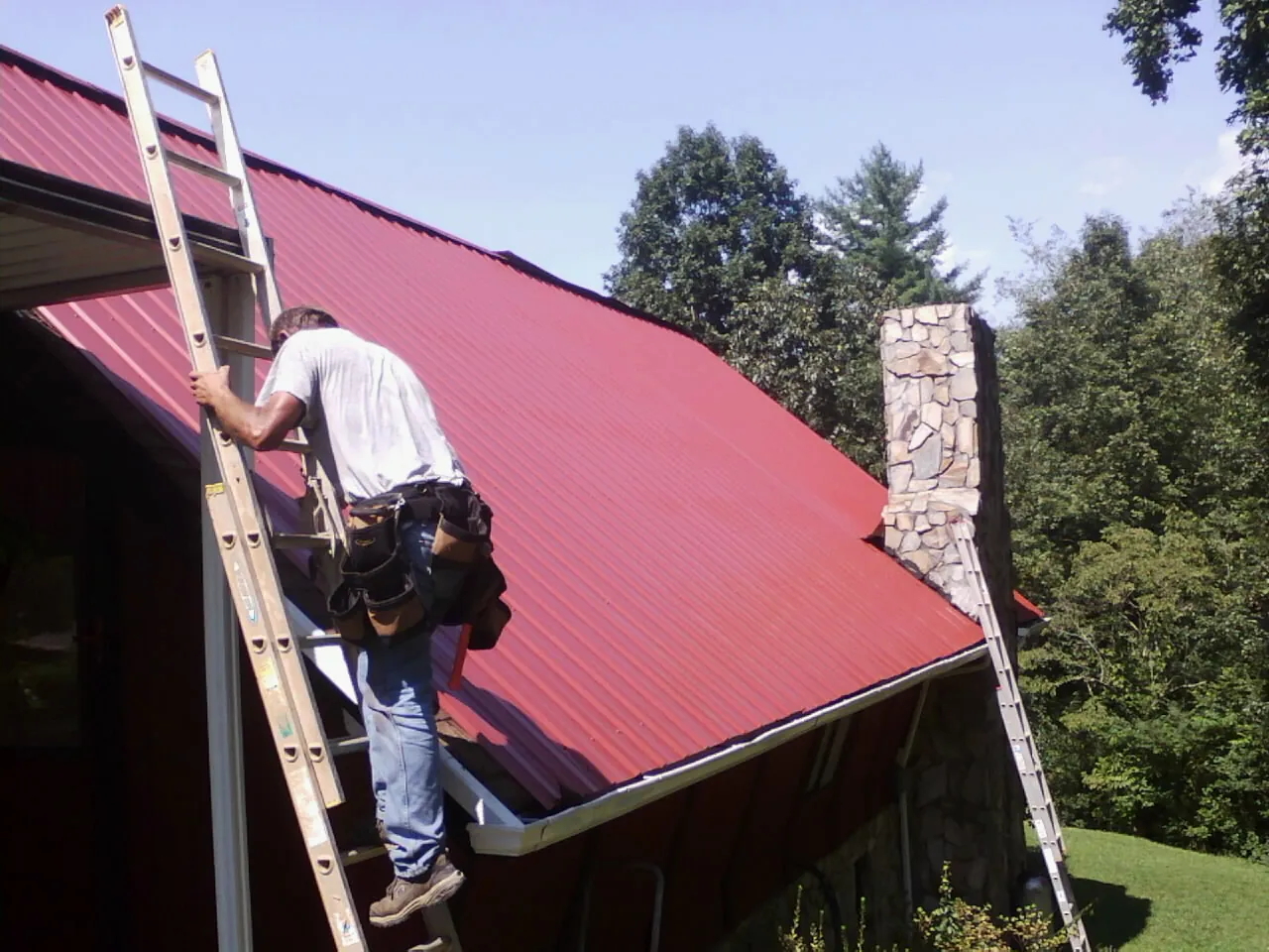 Roofer on a ladder beside a red metal roof and stone chimney — Soffit & Fascia Repair by Roofing Brothers in Beersheba Springs